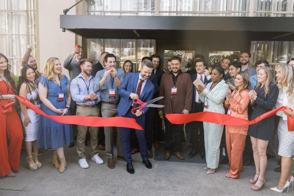 Mac Clemmens cutting a red ribbon with oversized scissors in front of an office building and a crowd of people